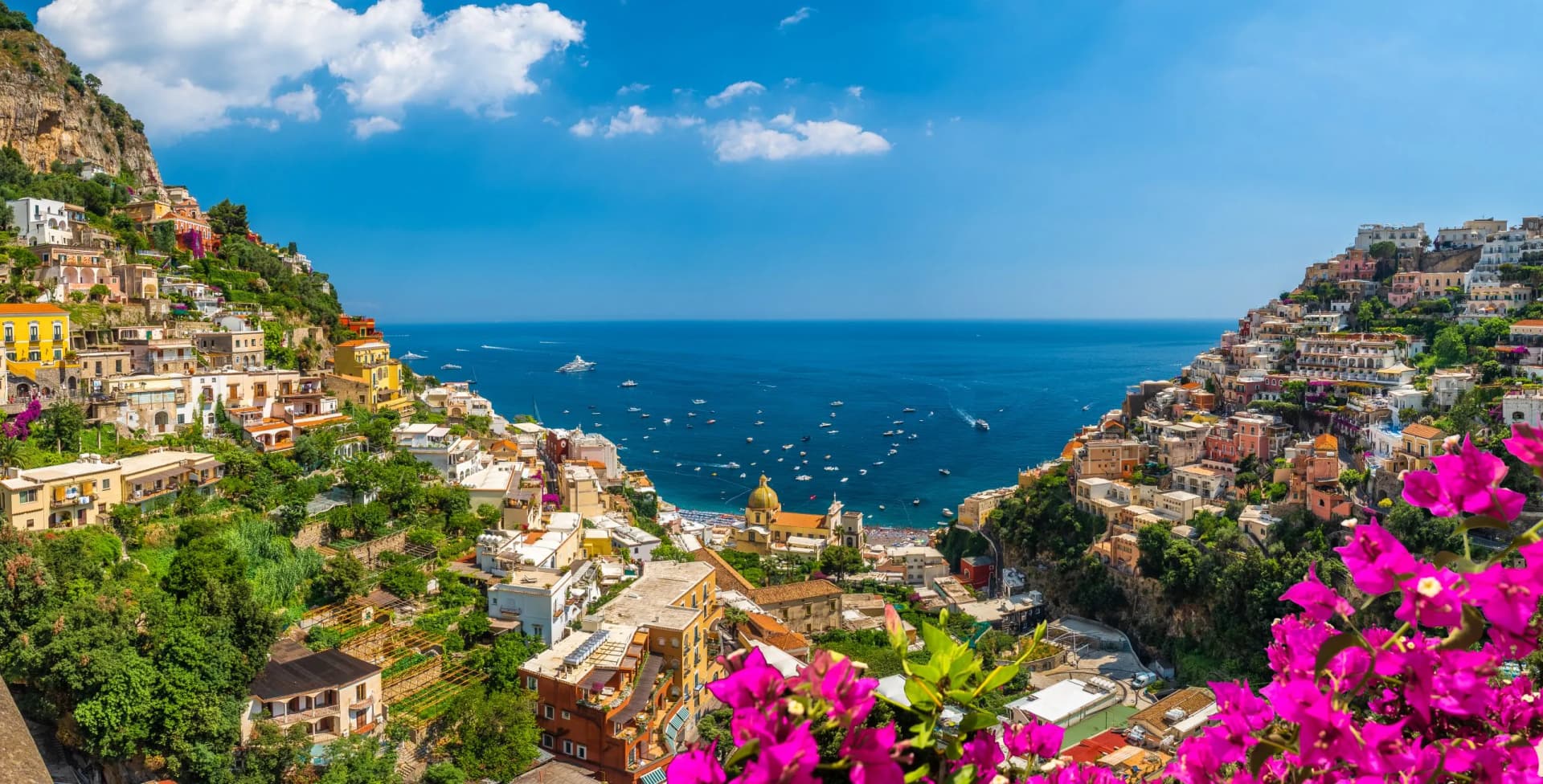 Coastal town built into steep cliffs overlooking the blue sea with many boats, bright pink flowers in foreground.